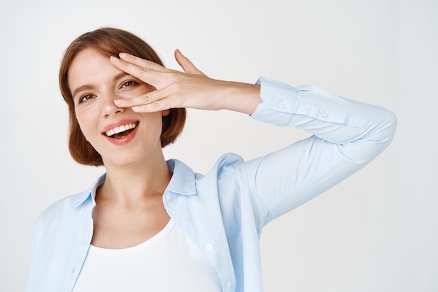 Portret van een gelukkige natuurlijke vrouw met kort haar die door de vingers kijkt met een dromerige, gefascineerde glimlach die op een witte achtergrond staat