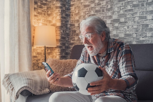 Portret van een gelukkige oude man die de overwinning van zijn liefdesteam van voetbal of voetbal op de bank viert Senior kijken naar een wedstrijd op zijn telefoon die een voetbalweddenschap wint