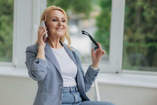 Portret van een gelukkige oude zakenvrouw in een bril die praat op een smartphone die de klant online raadpleegt, een glimlachende senior vrouw die spreekt op een mobiele telefoon, een draadloze verbinding gebruikt Technologieconcept