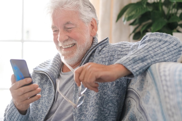 Portret van een gelukkige oudere man met een baard die ontspant op de bank thuis en naar een mobiele telefoon kijkt die lacht Headshot van een aantrekkelijke oudere man die geniet van zijn pensioen