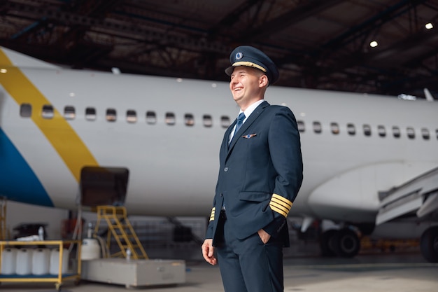 Portret van een gelukkige piloot in uniform die wegkijkt, klaar voor de vlucht, staande voor een groot passagiersvliegtuig in de luchthavenhangar. Vliegtuigen, beroep, transportconcept