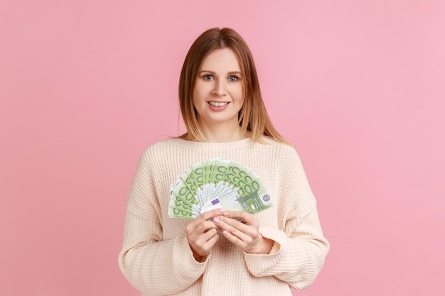 Portret van een gelukkige rijke blonde vrouw die een grote fan van eurobankbiljetten vasthoudt, naar de camera kijkt met een tevreden uitdrukking, een witte trui draagt. Indoor studio opname geïsoleerd op roze achtergrond.