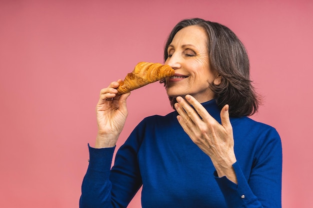 Portret van een gelukkige senior volwassen oude vrouw die croissant eet die over roze achtergrond wordt geïsoleerd. Frans ontbijtconcept.