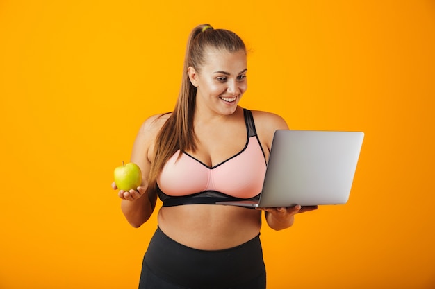 Portret van een gelukkige te zware jonge vrouw die sportkleding draagt die zich geïsoleerd over gele muur bevindt, die laptop computer gebruikt, die groene appel houdt