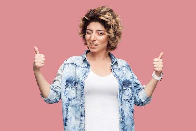 Portret van een gelukkige tevreden jonge vrouw met krullend kapsel in een casual blauw shirt dat staat, duimen omhoog, knipoogt en naar de camera kijkt met een brede glimlach. indoor studio opname, geïsoleerd op roze achtergrond.