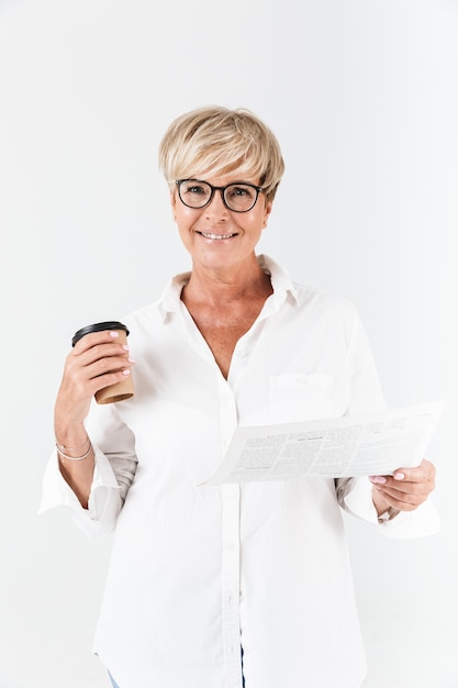 Portret van een gelukkige volwassen vrouw met een bril die een koffiekopje houdt en een krant leest die over een witte muur is geïsoleerd