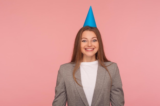 Portret van een gelukkige vrolijke vrouw in een pak en met een feestkegel op haar hoofd die lacht naar de camera, een verjaardag viert, een baanpromotie. indoor studio-opname geïsoleerd op roze achtergrond
