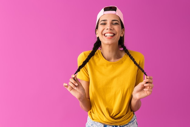 portret van een gelukkige, vrolijke vrouw met een pet achterstevoren glimlachend en aanrakend haar staartjes geïsoleerd op een roze muur