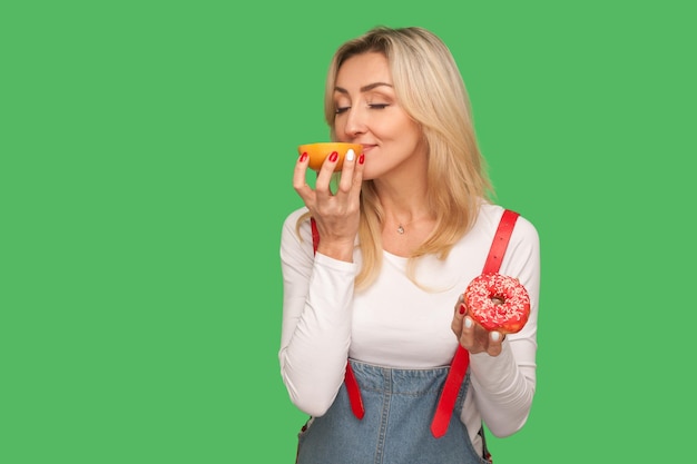 Portret van een gelukkige vrouw die geniet van de geur van heerlijke grapefruit terwijl ze zoete donut in de andere hand houdt, concept van het kiezen van gezond fruit in plaats van junkfood. studio-opname geïsoleerd op groene achtergrond