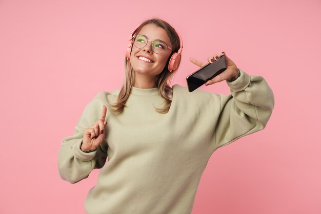 Portret van een gelukkige vrouw in een bril met een koptelefoon en een mobiele telefoon terwijl ze met de vinger naar boven wijst, geïsoleerd over roze muur