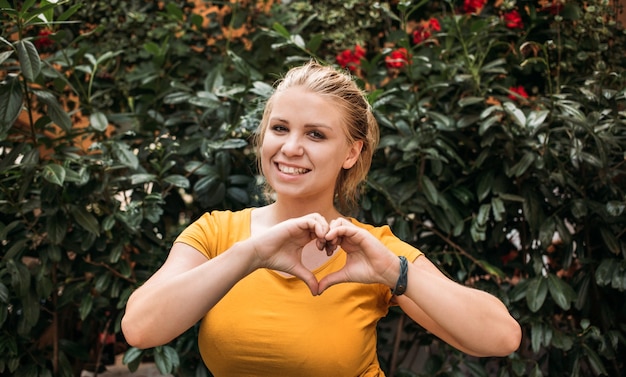 Portret van een gelukkige vrouw in een geel T-shirt in de tuin. Een hartvorm maken met haar handen