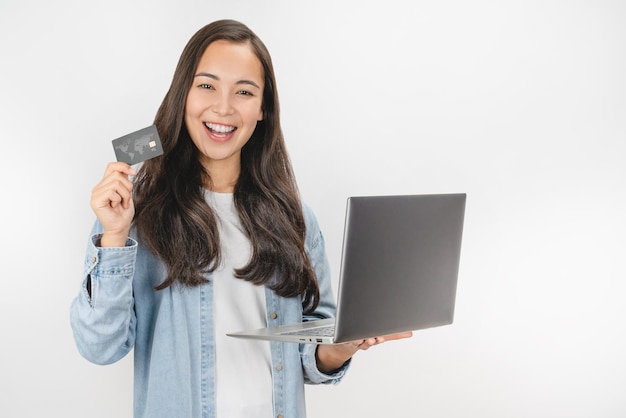Portret van een gelukkige vrouw in een spijkerbroek met een creditcard en een laptop die op een witte achtergrond wordt geïsoleerd