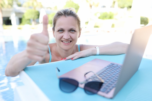 Portret van een gelukkige vrouw met een duim omhoog gebaar die op een laptop werkt terwijl ze in het zwembad op vakantie is