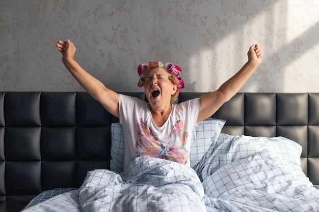 Portret van een gelukkige vrouw van middelbare leeftijd in krulspelden gegaap zittend op bed thuis slaapkamer. Luie middag concept.