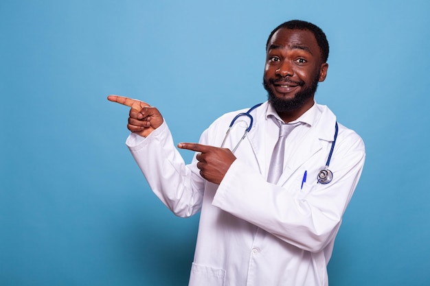 Portret van een gelukkige zelfverzekerde dokter met een witte laboratoriumjas en een stethoscoop die beide handen met wijsvingers naar zijn rechterkant wijst. Glimlachende arts die handgebaar doet die richting aangeeft.