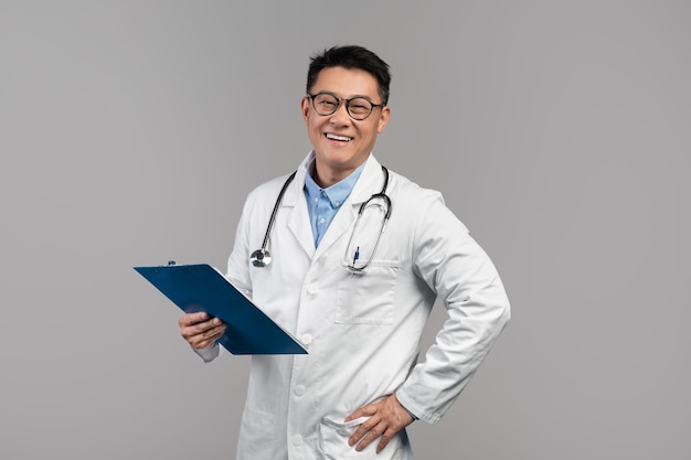 Portret van een gelukkige zelfverzekerde volwassen aziatische arts in een witte jasbril met stethoscoop en tablet kijkt naar de camera