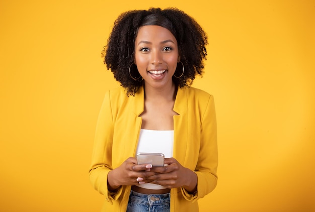 Portret van een gelukkige zwarte dame met borstelig haar die een mobiele telefoon gebruikt en naar de camera glimlacht