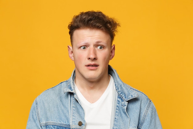 Portret van een geschokte, bange jonge man met denim vrijetijdskleding die opzij kijkt geïsoleerd op een geeloranje muurachtergrond in de studio. Mensen oprechte emoties, lifestyle concept. Bespotten kopie ruimte.