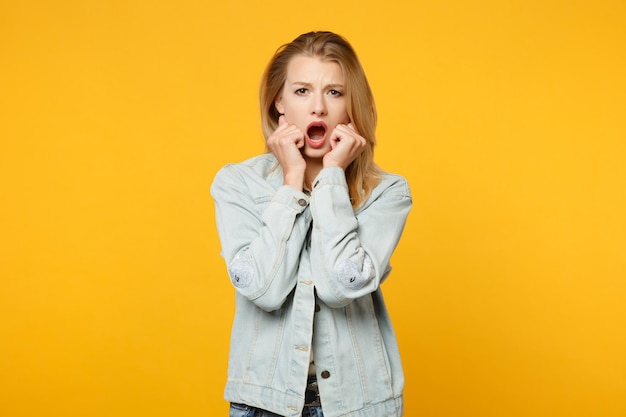 Portret van een geschokte, bange jonge vrouw in denim vrijetijdskleding die de mond openhoudt, handen op de wangen legt geïsoleerd op een geeloranje achtergrond in de studio. Mensen levensstijl concept. Bespotten kopie ruimte.