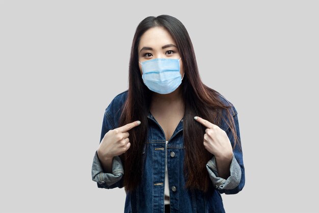 Portret van een geschokte brunette aziatische jonge vrouw met een chirurgisch medisch masker in een blauw spijkerjasje dat zichzelf wijst en naar de camera kijkt. indoor studio opname, geïsoleerd op een grijze achtergrond.