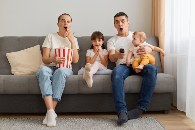 Portret van een geschokte familie die binnenshuis mier thuis op de bank zit en tv of horrorfilm kijkt die schok en verrassing uitdrukt in casual kleding die popcorn eet