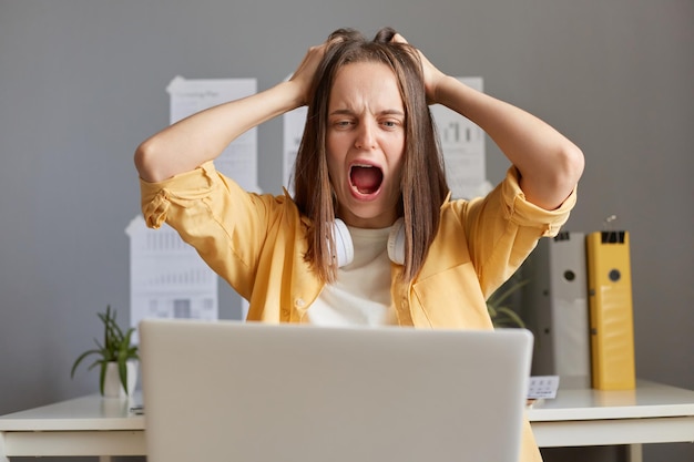 Portret van een geschokte, gestresste depressieve vrouw met bruin haar die op een laptop werkt en grote problemen heeft, niet weet wat ze moet doen, luid schreeuwend en aan haar haar trekkende achtergrond