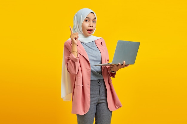 Portret van een geschokte jonge Aziatische vrouw die een laptop vasthoudt en omhoog wijst, geïsoleerd op een gele achtergrond