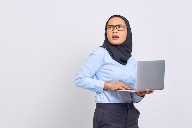 Portret van een geschokte jonge Aziatische vrouw die een laptop vasthoudt en wegkijkt op een witte achtergrond