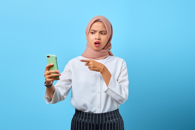 Portret van een geschokte jonge Aziatische vrouw die op een mobiele telefoon wijst met open mond over een blauwe achtergrond