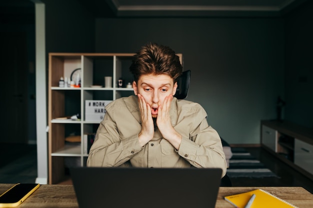 Portret van een geschokte jonge freelancer-man die op een laptop thuis werkt en naar het scherm kijkt met een boos gezicht verstoorde werknemer in een shirt werkt thuis op een computer quarantainewerk op afstand