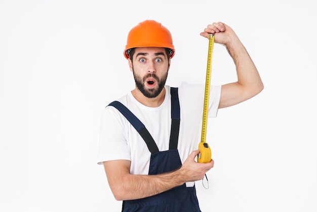 Portret van een geschokte jonge man bouwer in helm poseren geïsoleerd over witte muur met centimeter.