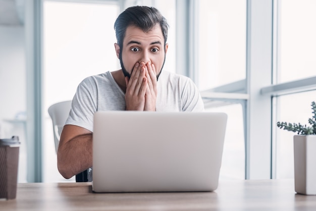 Portret van een geschokte jonge man die op een laptop werkt en zijn mond bedekt met handen