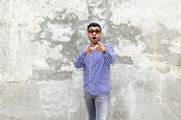 Portret van een geschokte knappe bebaarde jongeman in een geruit blauw shirt en een zonnebril die tegen een betonnen grijze muur staat, wijzend en kijkend naar de camera met een geschokt ongelooflijk gezicht.