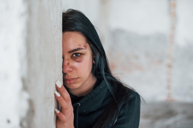 Portret van een geslagen jonge vrouw met blauwe plek onder het oog die binnenshuis in een verlaten gebouw op de muur staat en leunt