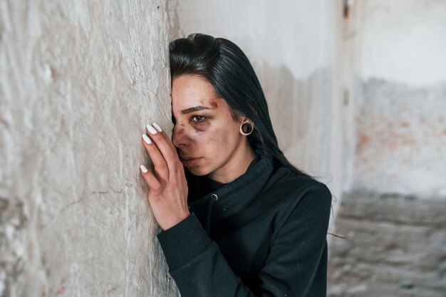 Portret van een geslagen jonge vrouw met blauwe plek onder het oog die binnenshuis in een verlaten gebouw op de muur staat en leunt