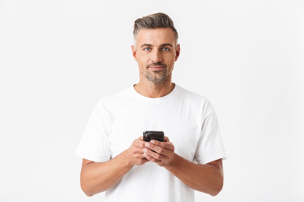 Portret van een gespierde man van 30 jaar die een casual t-shirt draagt en een smartphone gebruikt die op wit wordt geïsoleerd