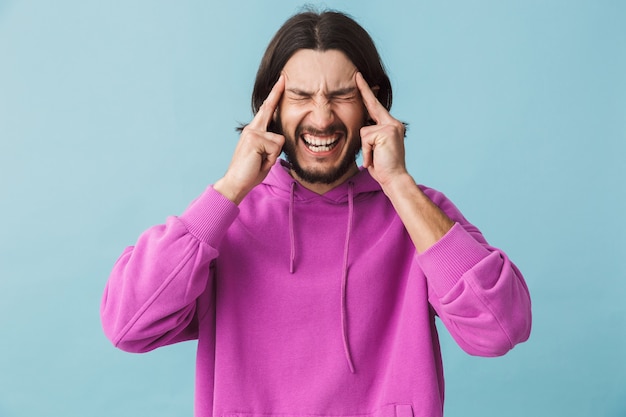 Portret van een gestresste jonge, bebaarde brunette man met een hoodie die geïsoleerd over een blauwe muur staat en hoofdpijn heeft