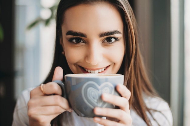 Portret van een geweldig Kaukasisch meisje kijken camera glimlachen terwijl in de buurt van haar lippen een kop warme koffie in een coffeeshop close-up.