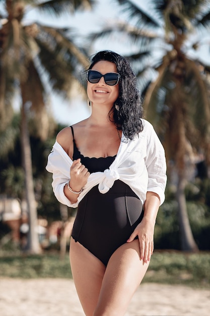 Portret van een geweldige jonge vrouw met een charmante sneeuwwitte glimlach in zonnebril poseren op het strand tegen de achtergrond van een palmboom. Vakantieconcept.