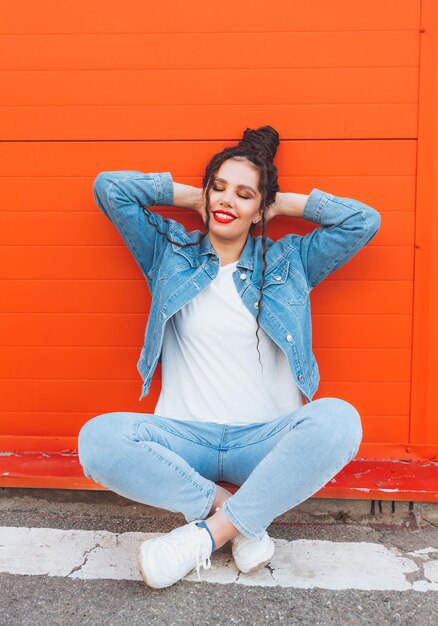 Portret van een glamoureuze vrouw met dreadlocks in een spijkerpak en met rode lippen tegen een oranje muur