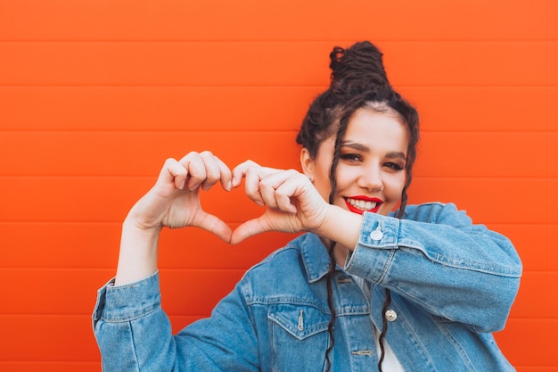 Portret van een glamoureuze vrouw met dreadlocks in een spijkerpak en met rode lippen tegen een oranje muur