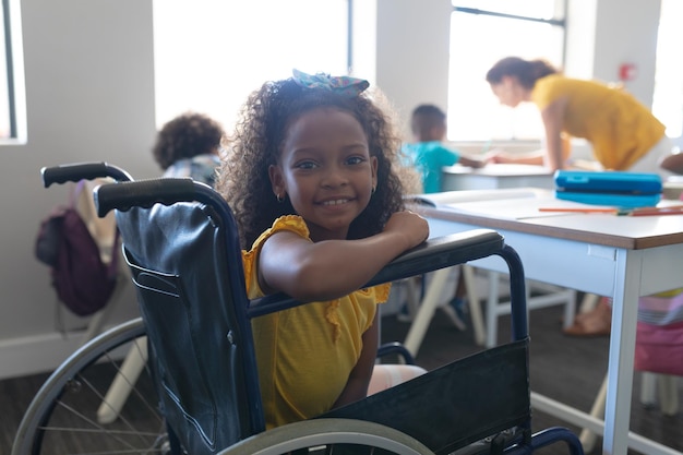 Portret van een glimlachend Afro-Amerikaans basisschoolmeisje dat op een rolstoel zit aan een bureau in de klas