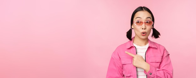 Portret van een glimlachend Aziatisch meisje in een stijlvolle outfitzonnebril die met de vinger naar links wijst en een advertentiebanner toont die over een roze achtergrond staat