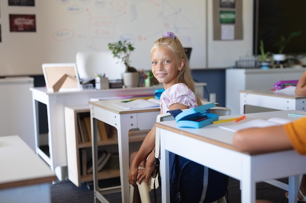 Foto portret van een glimlachend blank basisschoolmeisje dat aan een bureau in de klas zit