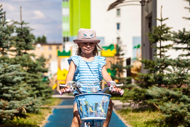 Portret van een glimlachend gelukkig klein meisje in een zomerhoed die buiten fietst in het stadspark en naar de camera kijkt