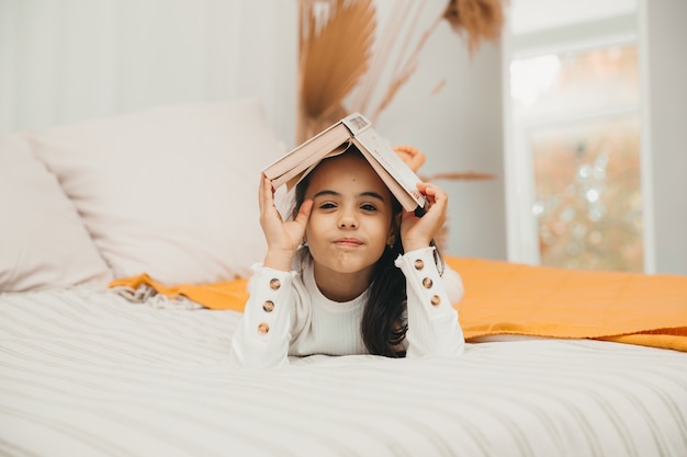 Portret van een glimlachend gelukkig meisje dat een boek op haar hoofd houdt en op het bed ligt.