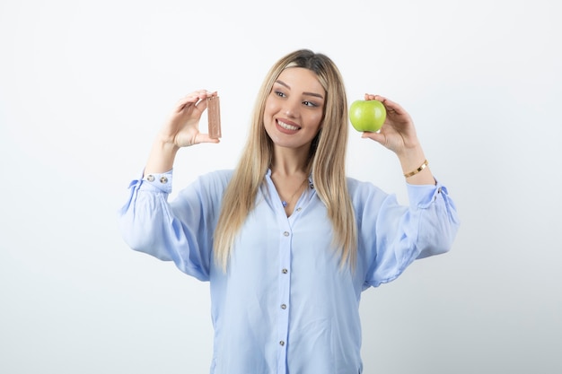 Portret van een glimlachend gelukkig meisje dat groene appel met chocoladereep houdt tegen witte muur