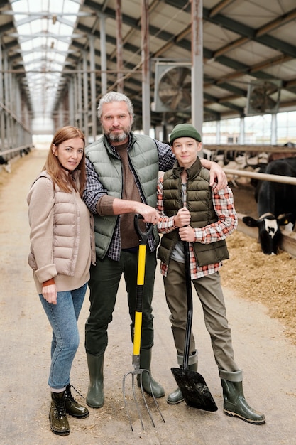 Portret van een glimlachend gezin met tienerzoon die samenwerkt op een veehouderij, vader die zoon omhelst en hooivork vasthoudt