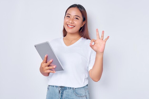 Portret van een glimlachend jong Aziatisch meisje in een wit t-shirt met een digitale tablet met een goed teken dat op een witte achtergrond wordt geïsoleerd