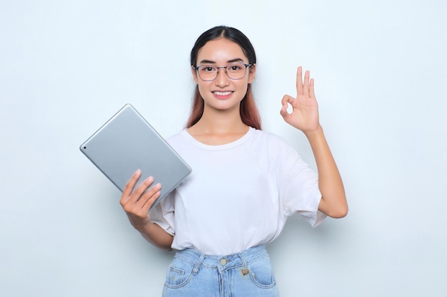 Portret van een glimlachend jong Aziatisch meisje in een wit t-shirt met een digitale tablet met een goed teken dat op een witte achtergrond wordt geïsoleerd